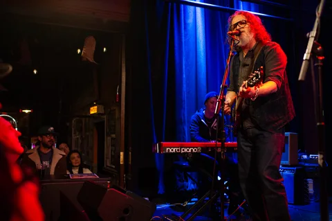 Guitarist at a microphone beside the blue stage curtain at the 2025 Jason Molina MusiCares benefit.