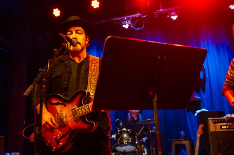 Singer wearing a hat, playing guitar at a microphone at the 2025 Jason Molina MusiCares benefit.