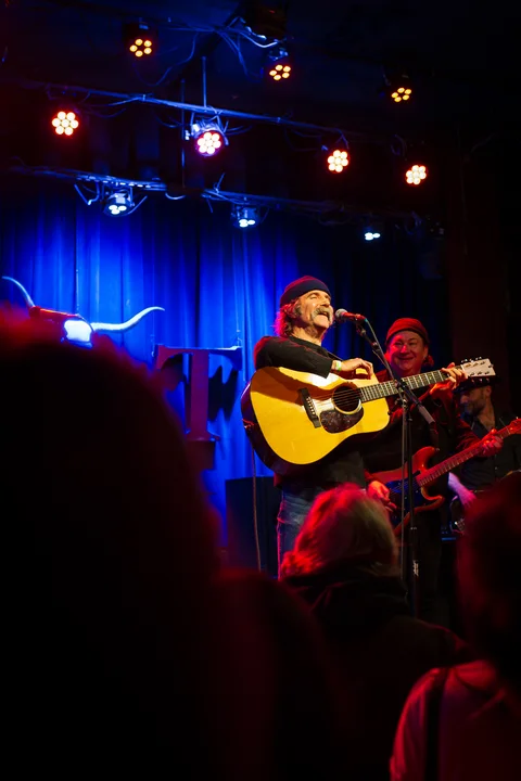 Acoustic guitarist at a microphone, seen from the crowd at the 2025 Jason Molina MusiCares benefit.