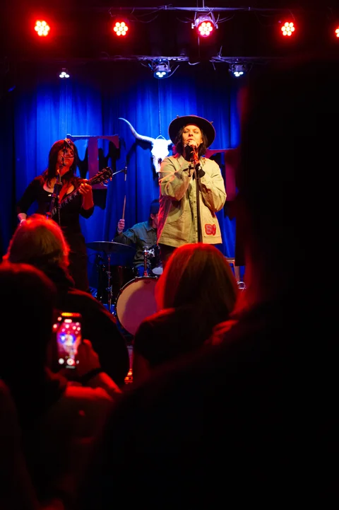 Two performers at microphones, seen from the audience at the 2025 Jason Molina MusiCares benefit.