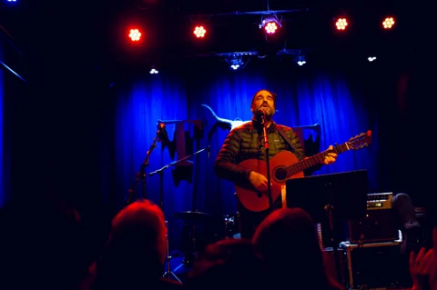 Singer and electric guitarist under the stage lights at the 2025 Jason Molina MusiCares benefit.