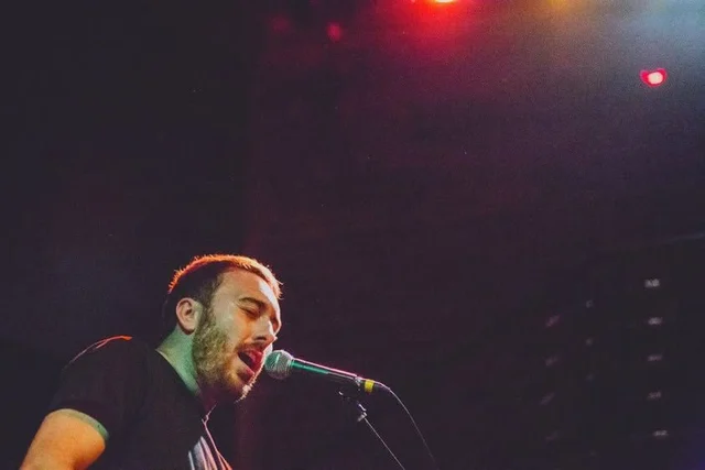 Performer at a microphone beneath colored stage lights. The Trouble Starts, East release show, August 16, 2013.
