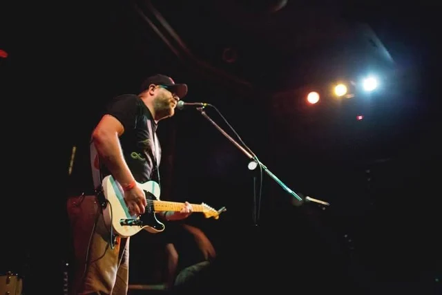 Guitarist at a microphone under overhead stage lights. The Trouble Starts, East release show, August 16, 2013.