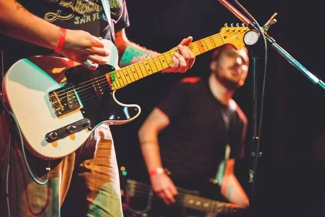 Cream electric guitar in the foreground with a band member behind. The Trouble Starts, East release show, August 16, 2013.