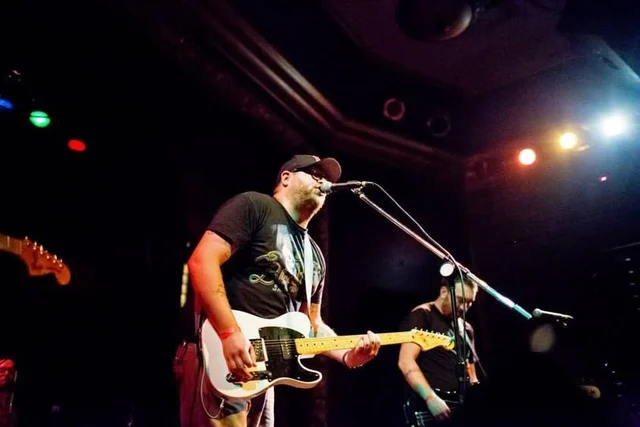 Guitarist playing a cream electric guitar at a microphone. The Trouble Starts, East release show, August 16, 2013.