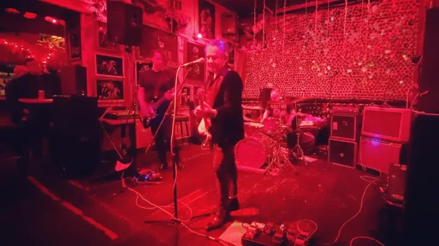 Graveyard of the Pacific performing at Cha Cha Lounge under red lighting, with a guitarist at the microphone, a bassist, and a drummer.