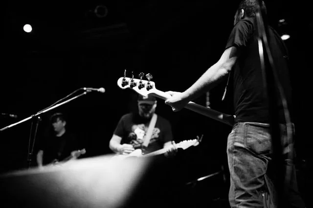 Black-and-white view across the stage toward a guitarist. The Trouble Starts, East release show, August 16, 2013.